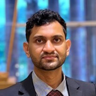 Man with short curly hair and beard wearing a dark suit, grey shirt, and maroon tie.