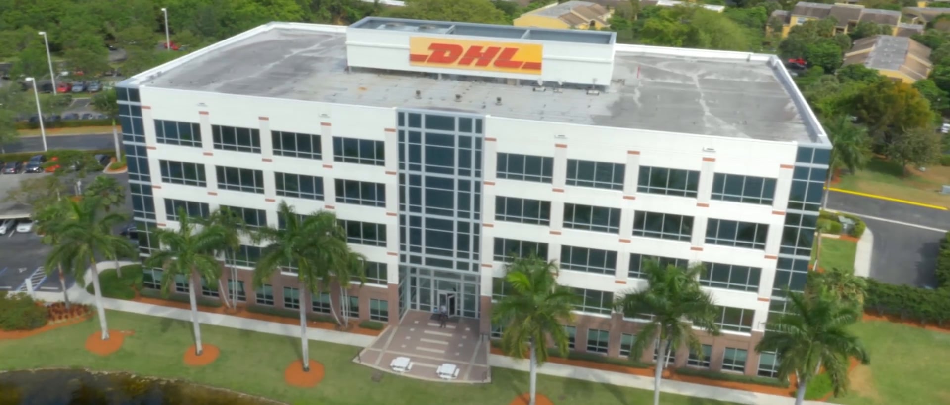 Four-story DHL office building with glass windows, palm trees, and parking lot surrounding it.