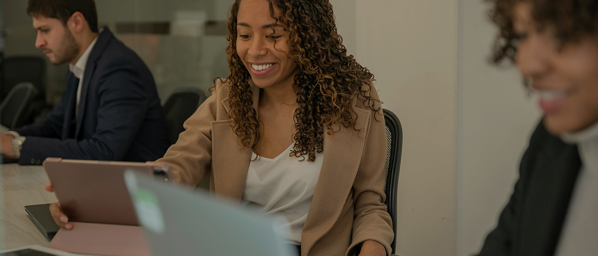 Smiling woman with curly hair using a tablet in an office with colleagues working nearby.