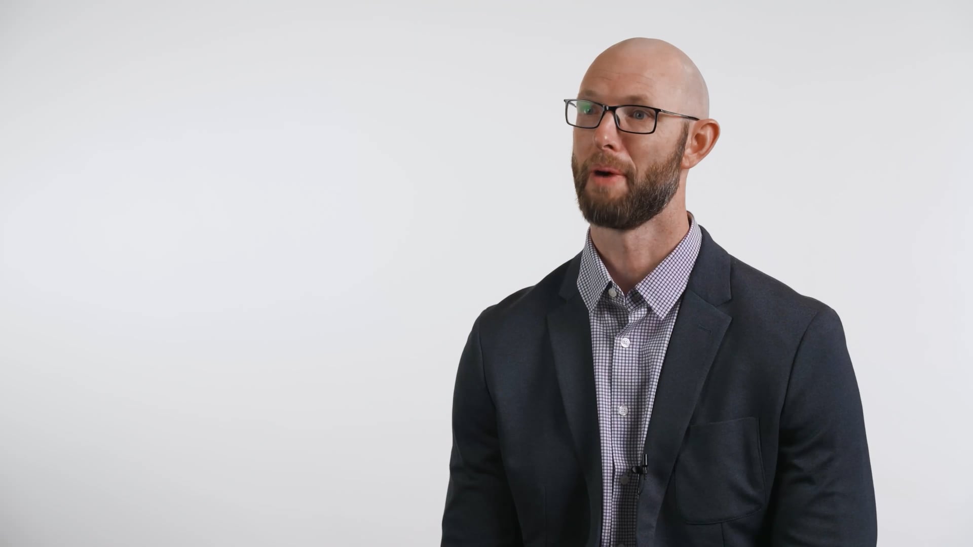 Bald man with glasses and beard wearing a checkered shirt and dark blazer against a white background.