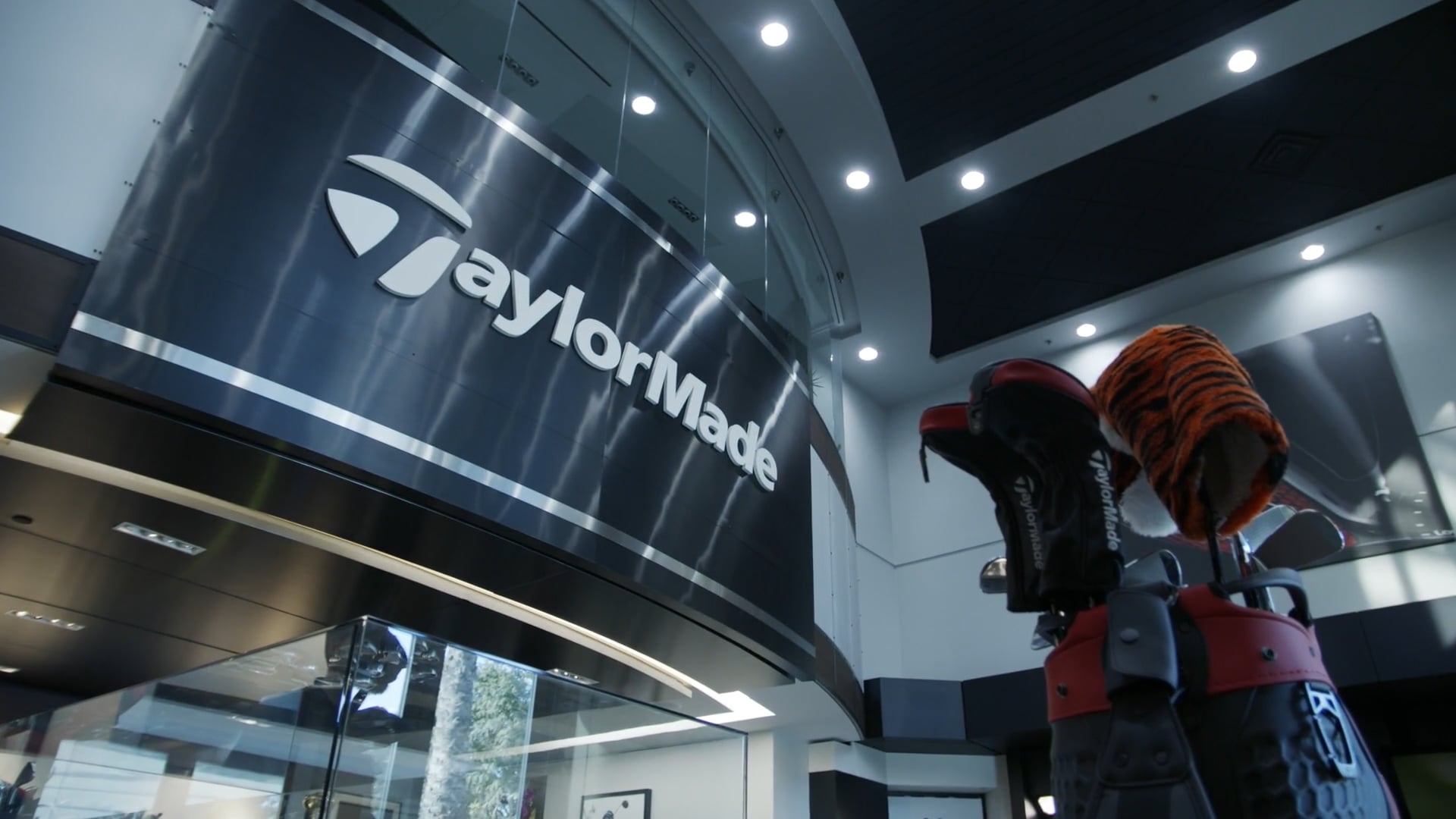 Indoor view of TaylorMade logo on a curved wall with a golf bag and clubs in the foreground.