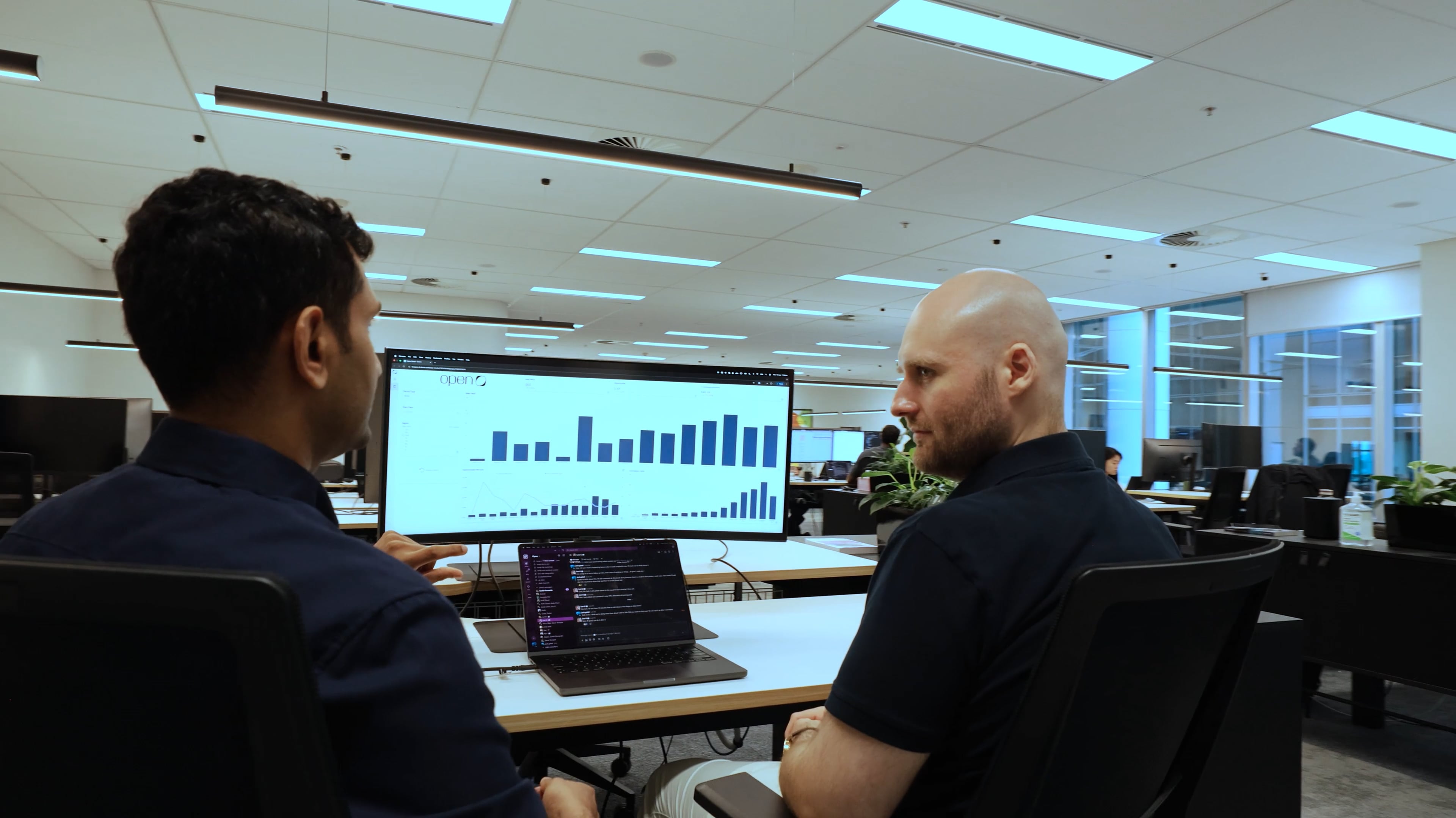 Two men in an office discuss bar graphs displayed on a large monitor and laptop screen.