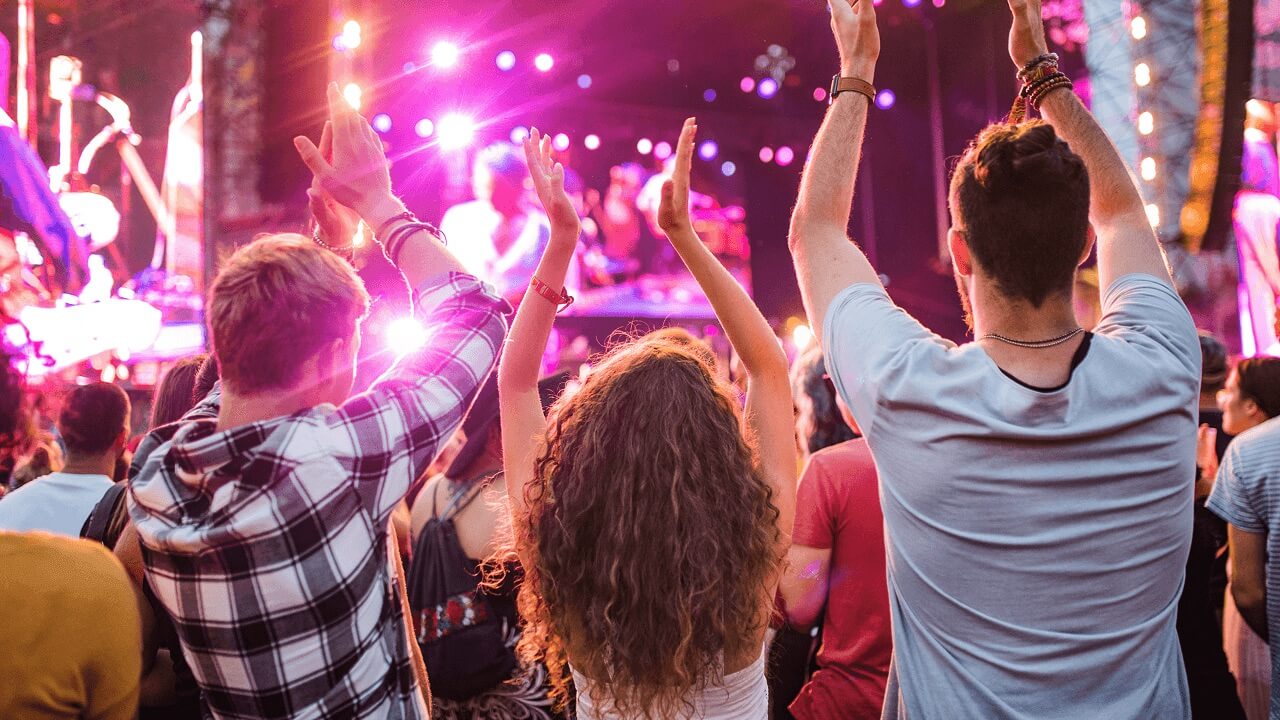 Crowd at a concert with people raising hands towards a brightly lit stage with purple and pink lights.