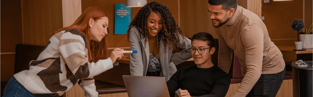 Four young professionals gathered around a laptop, discussing and smiling in a cozy office space.