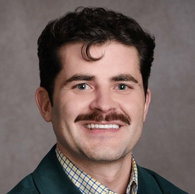 Portrait of a man with dark curly hair, mustache, and checkered shirt under a dark jacket.
