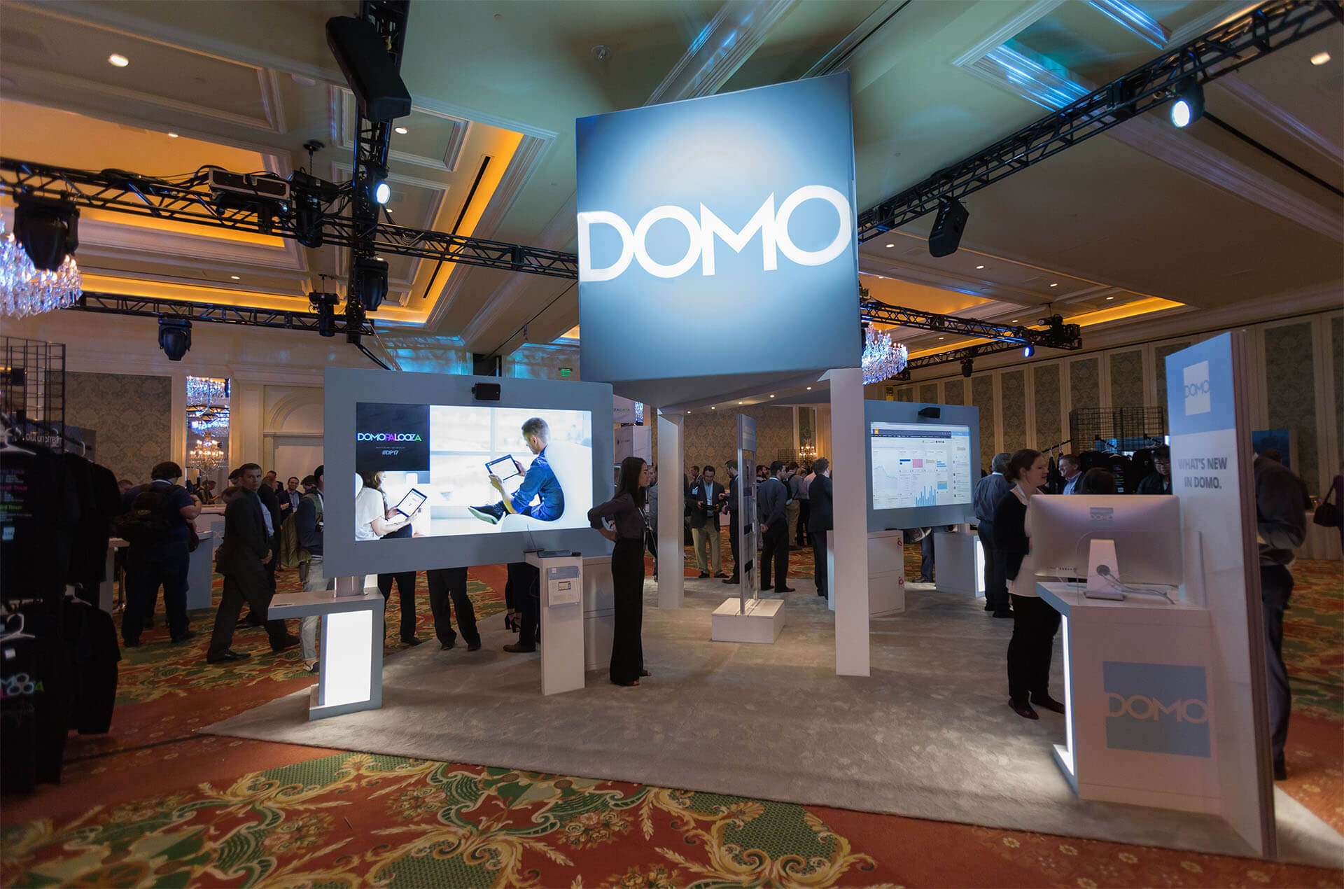 Trade show booth with large DOMO sign, display screens, and attendees in a conference hall.