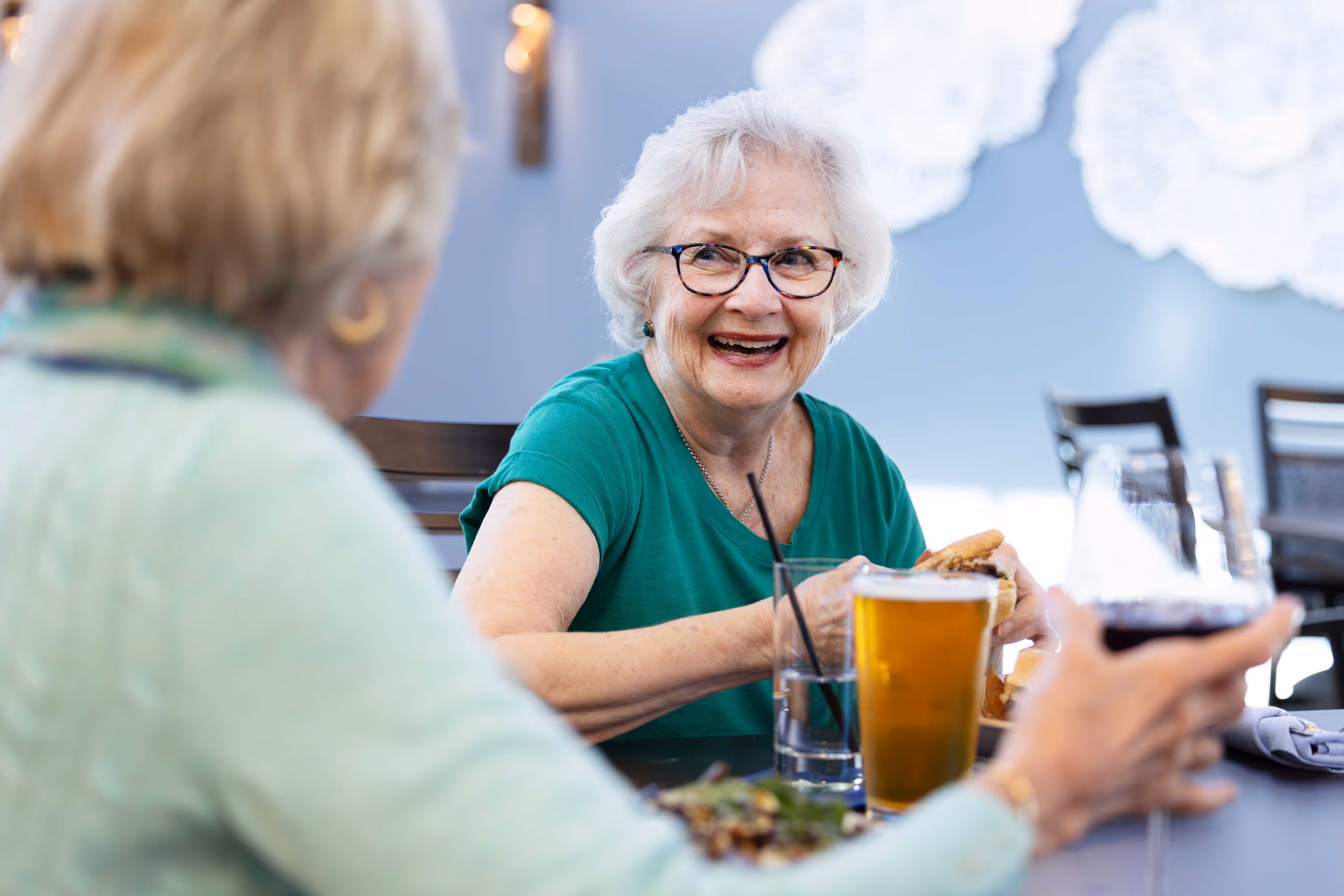 Older woman smiling and enjoying a meal with a beer, sitting with a friend in a relaxed, cheerful setting.