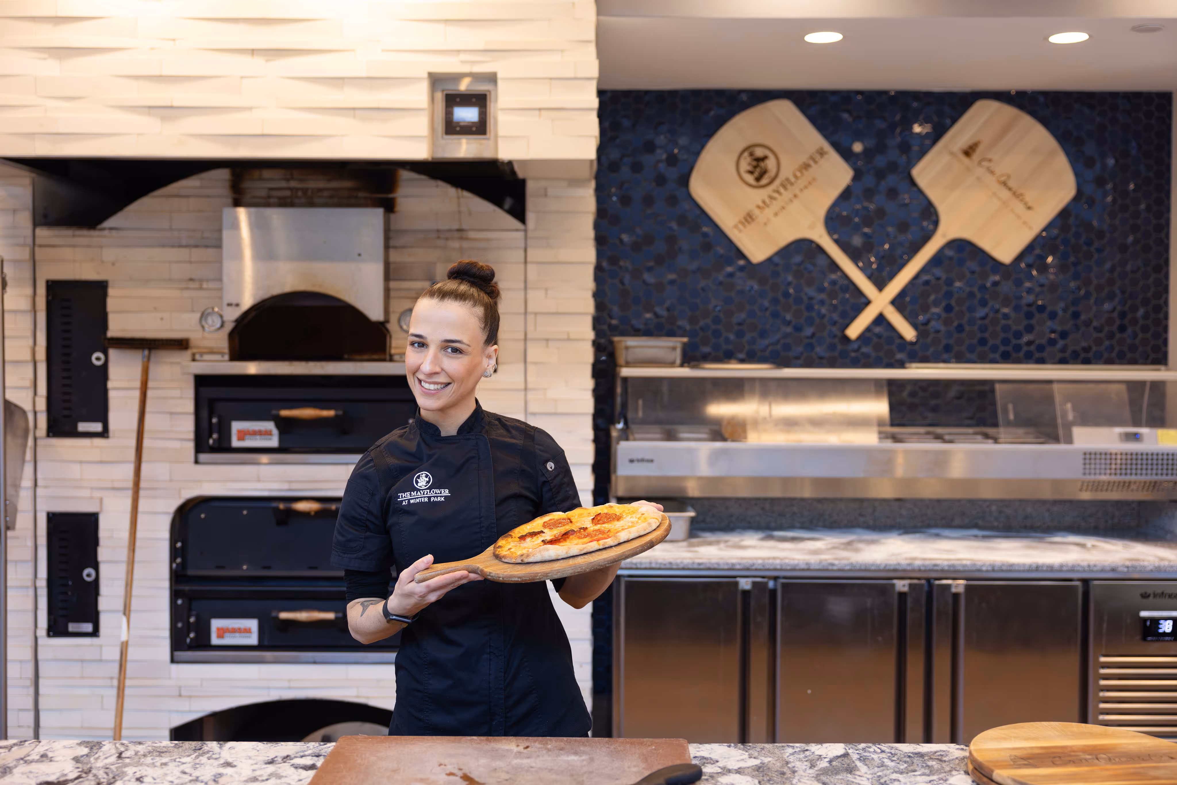 Chef proudly holding a freshly made pizza in front of a stone oven, showcasing their culinary creation.