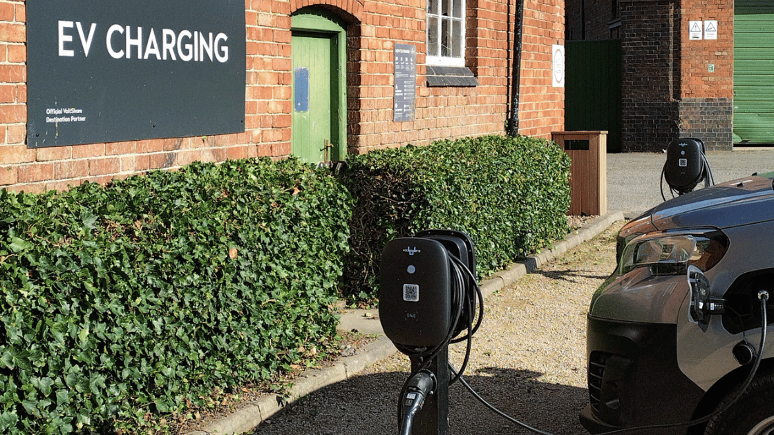 Electric car plugged into a voltshare charger outside a brick building with an EV charging sign.