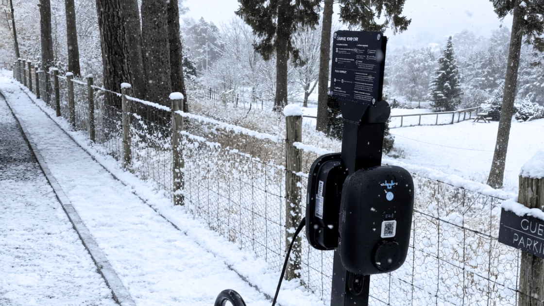 Black EV charger beside a snow-covered path and fence in a rural setting