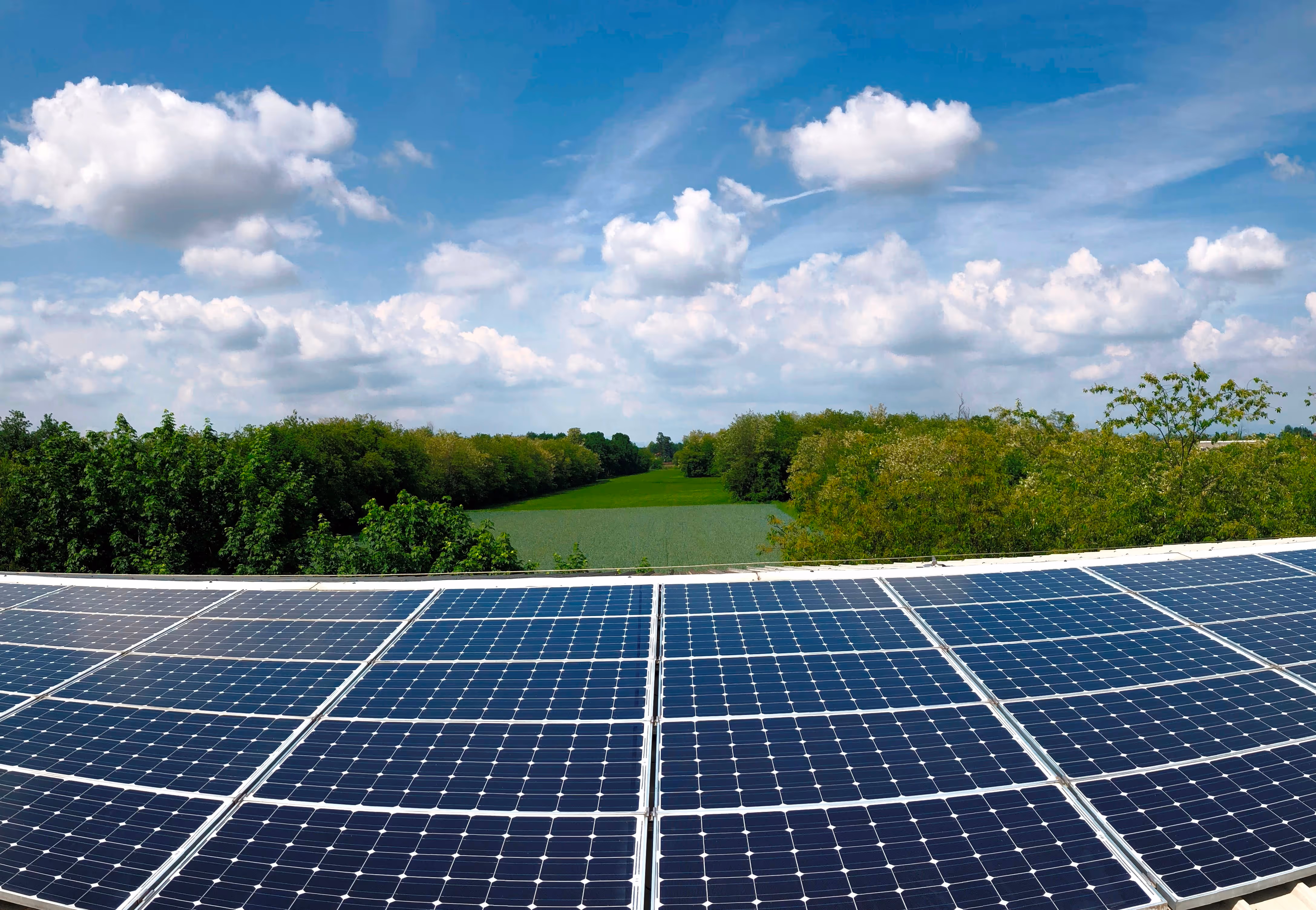 Pannelli solari sul tetto con un campo verde e alberi sotto un cielo azzurro con nuvole bianche.