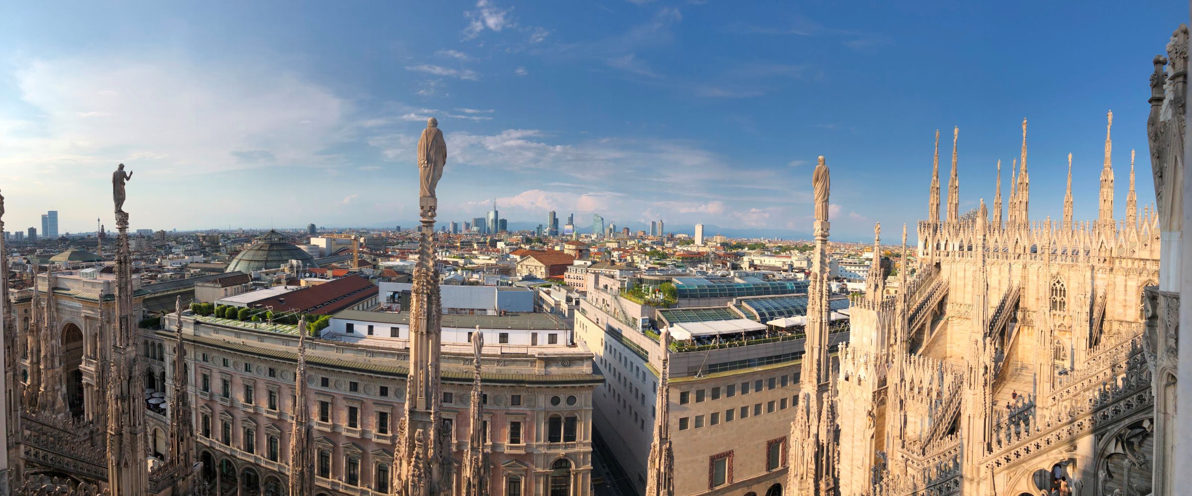 Panoramica della città di Milano con le guglie e le statue del Duomo e lo skyline moderno sullo sfondo sotto un cielo sereno.