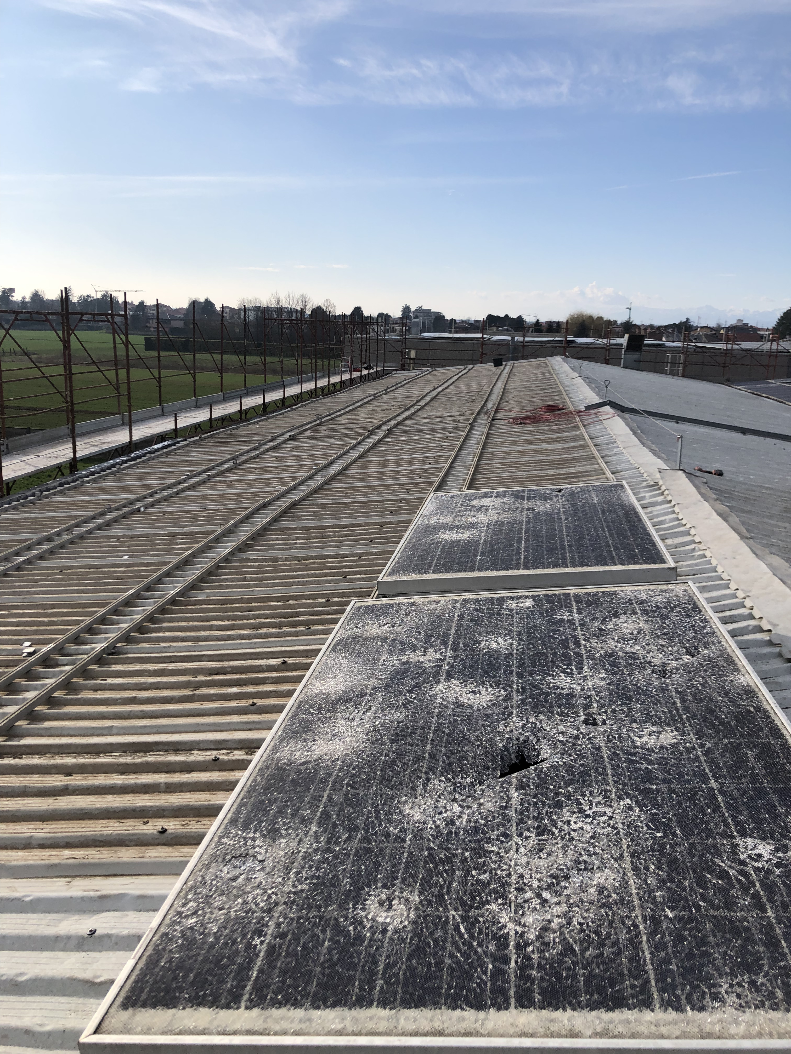 Due pannelli solari sporchi e danneggiati installati su un tetto in metallo all'aperto con un cielo azzurro e un paesaggio con campi verdi sullo sfondo.