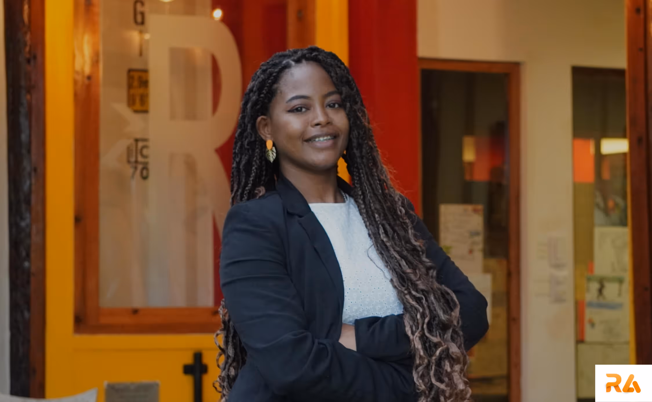 A lawyer with long braided hair wearing a black blazer and white blouse stands with arms crossed, smiling in front of a lawyer office.