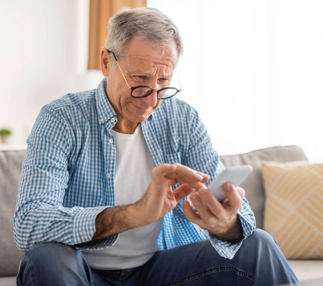 Older man with gray hair and glasses sitting on a couch, looking closely at a smartphone with a concerned expression, suggesting difficulty seeing due to macular degeneration.