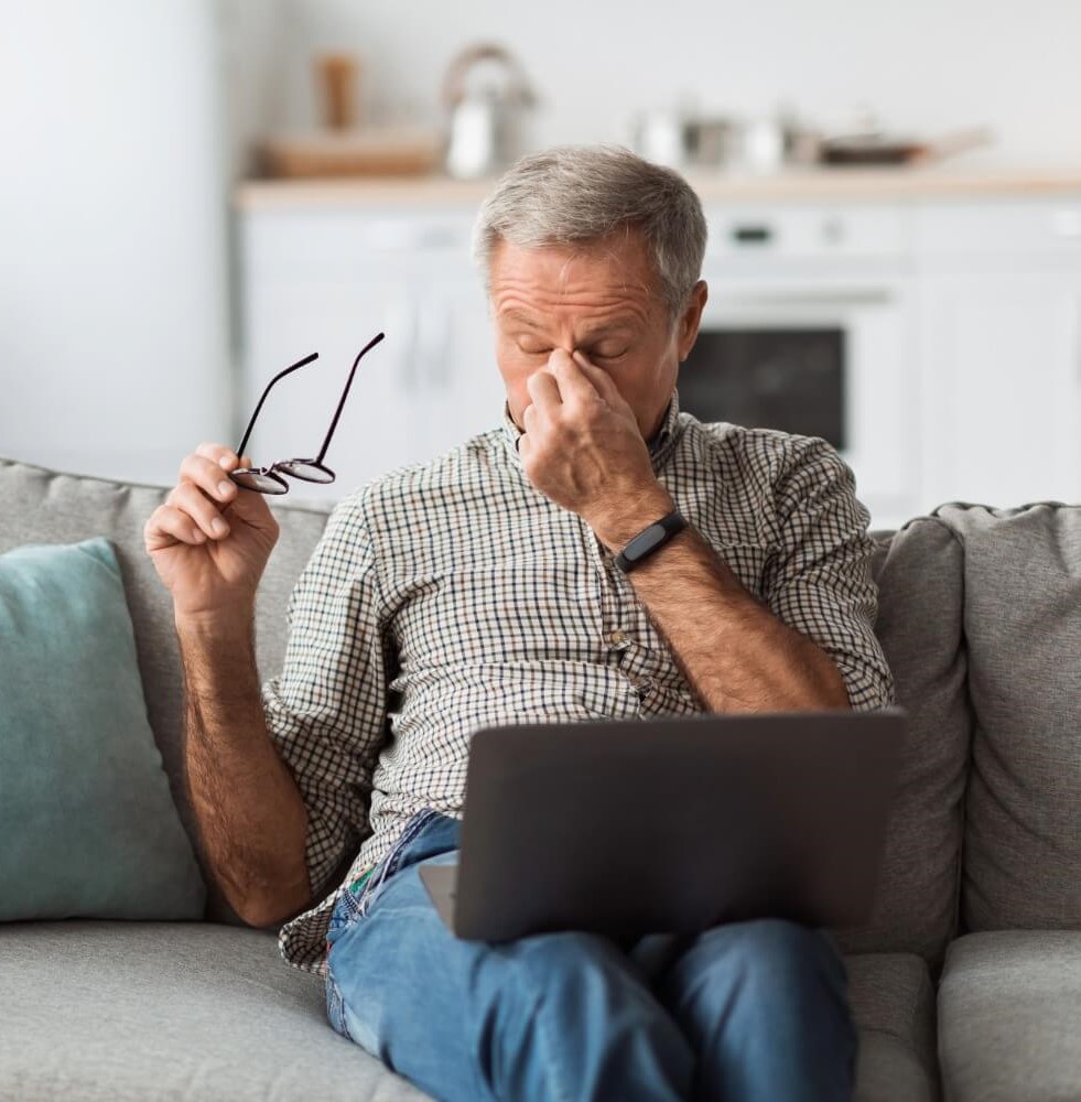 Older man sitting on a couch with a laptop on his lap, holding his glasses and pinching the bridge of his nose as if experiencing eye strain or vision problems related to macular degeneration.