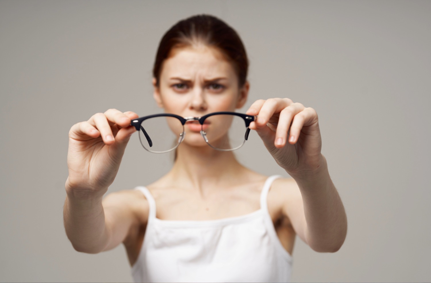 Woman in white top holding eyeglasses towards the camera with a focused expression.
