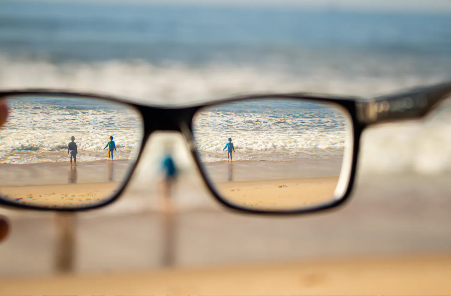 View of two children standing on a beach near ocean waves seen clearly through a pair of eyeglasses, while the surrounding beach is blurred.