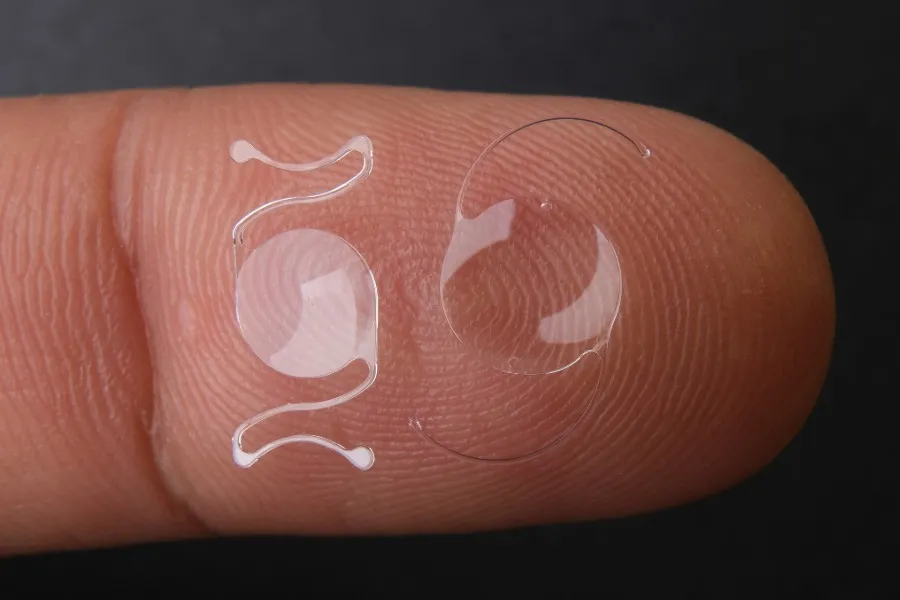 Close-up of two clear intraocular lenses resting on the tip of a finger against a dark background.