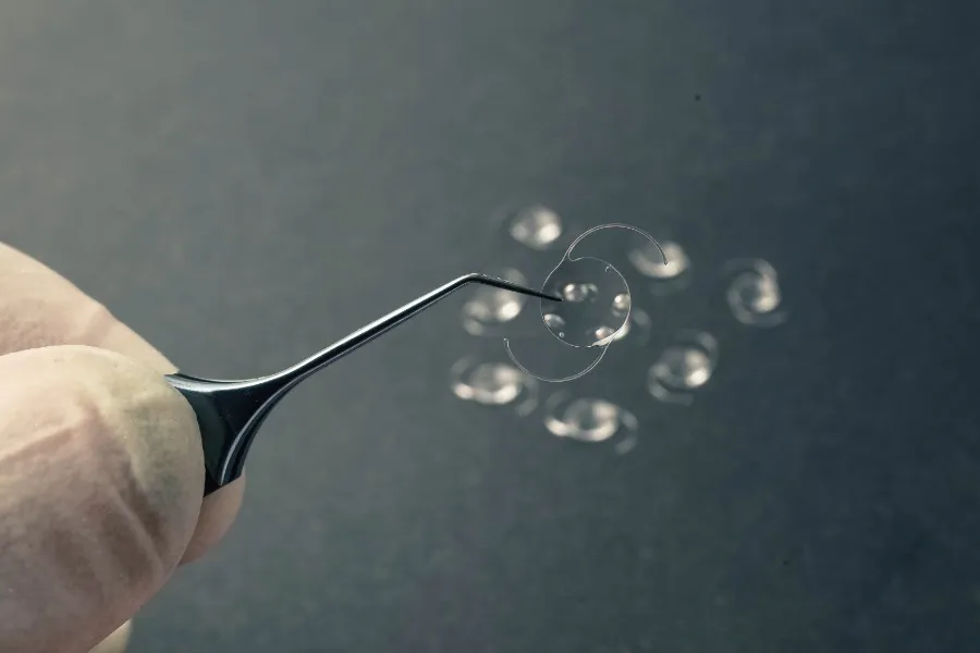 Close-up of a hand holding tweezers with a transparent toric contact lens with multiple lenses blurred in the background.