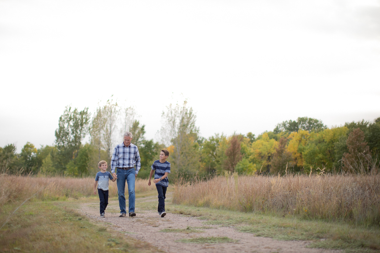 Older man walking outdoors on a dirt path holding hands with a young boy, accompanied by another boy walking beside them.