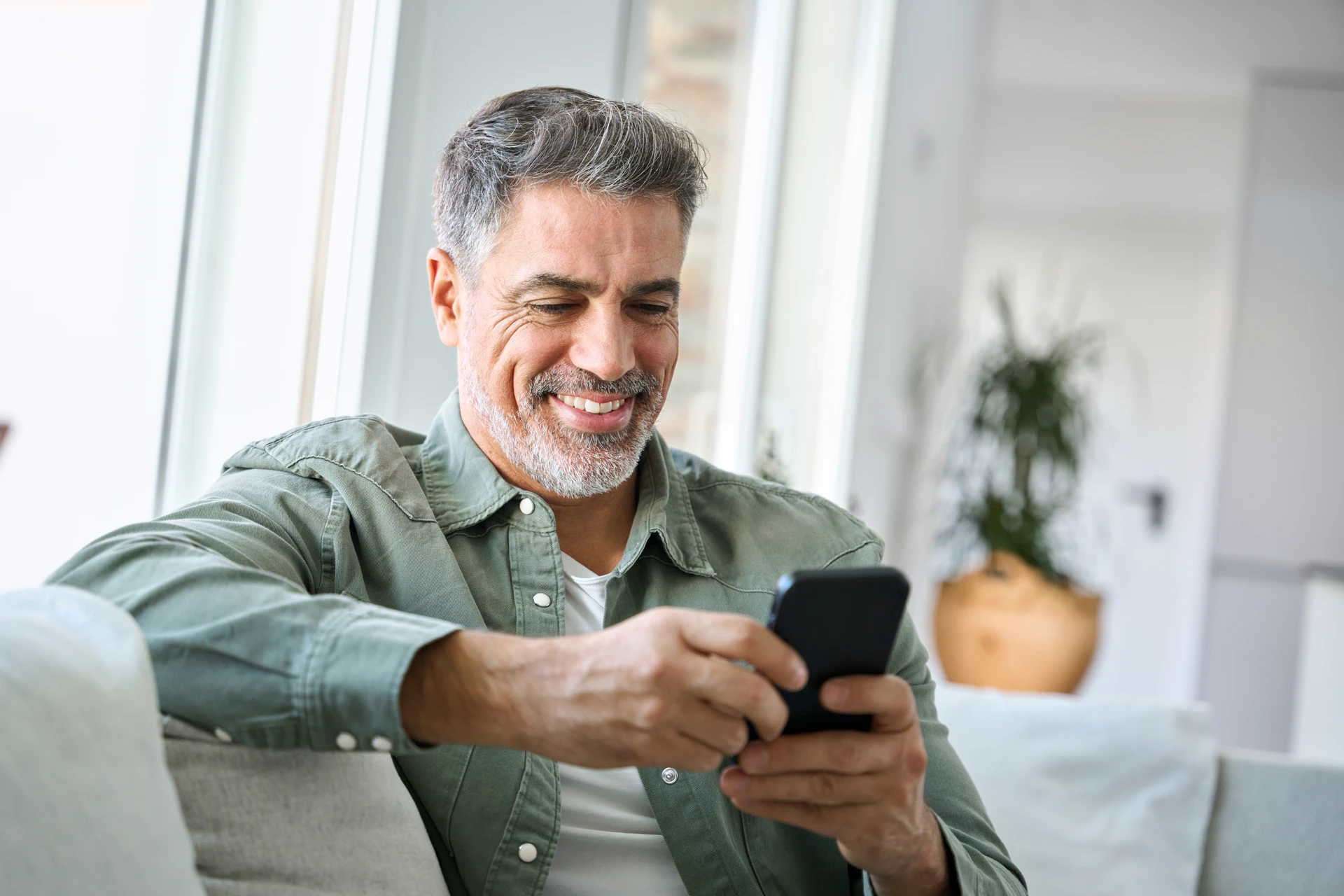 Smiling middle-aged man sitting on a couch and looking at his smartphone in a bright living room.