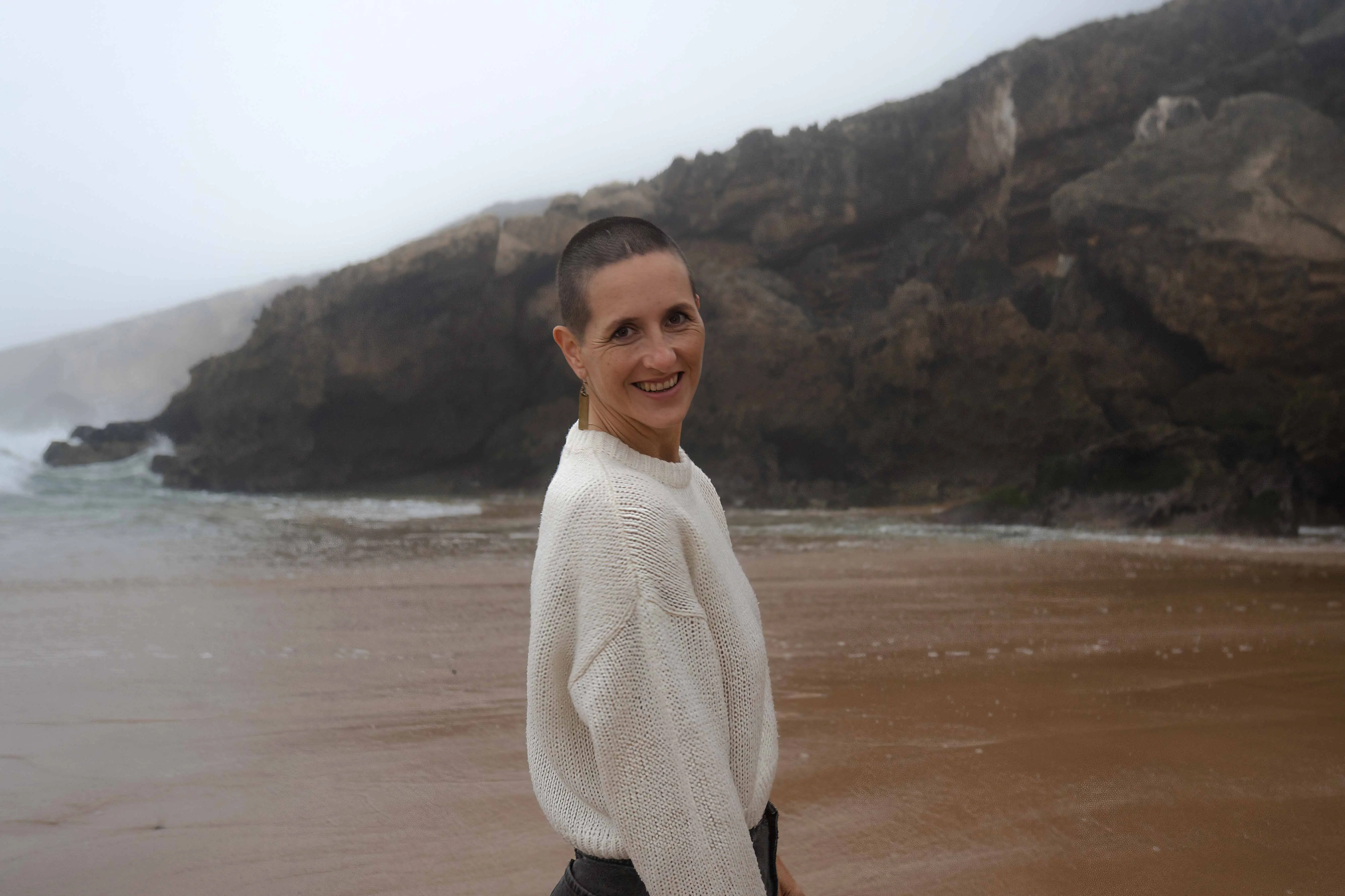 Rahel stands barefoot on a quiet beach with rocky cliffs and ocean waves behind her, turning toward the camera with a relaxed smile. The open coastal landscape reflects the grounded and human presence behind her work in somatic astrology.