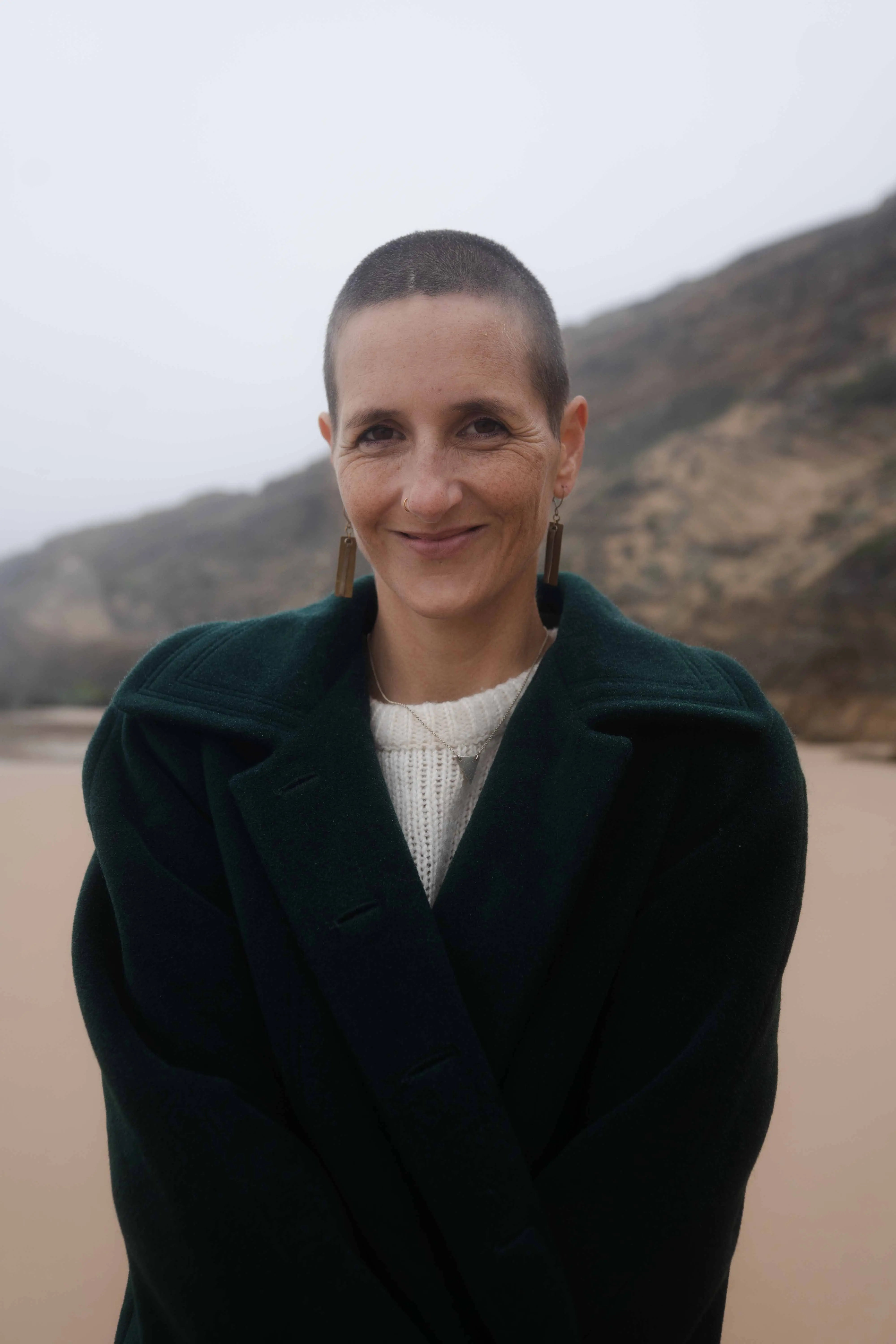 ahel stands on a misty beach with coastal cliffs rising behind her, wrapped in a dark coat and offering a soft welcoming smile. The calm presence reflects the supportive space of a somatic astrology mentorship and invites women to begin with an introductory Ashray call.