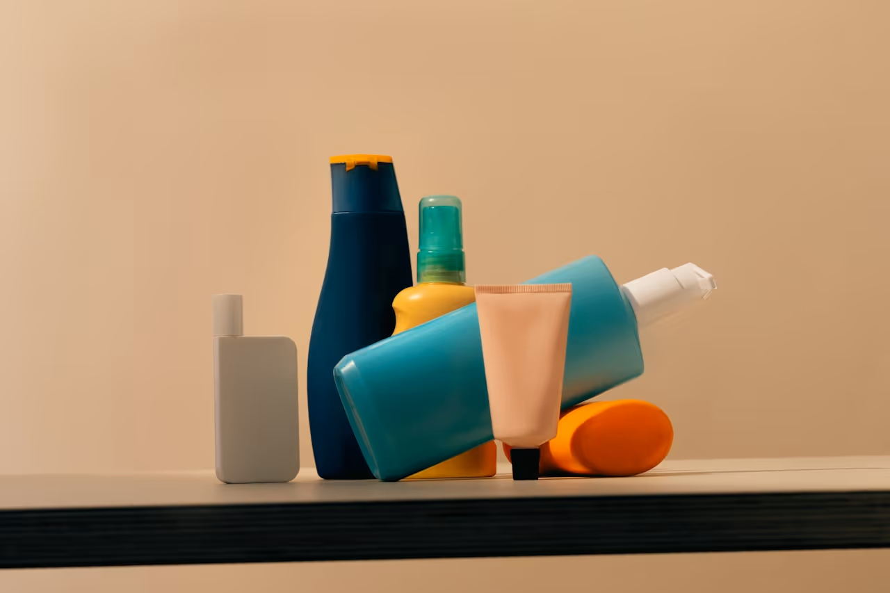 A collection of colorful cosmetic and personal care bottles, including a blue shampoo bottle, a yellow spray, and a tube of lotion, arranged on a wooden shelf against a soft beige background.