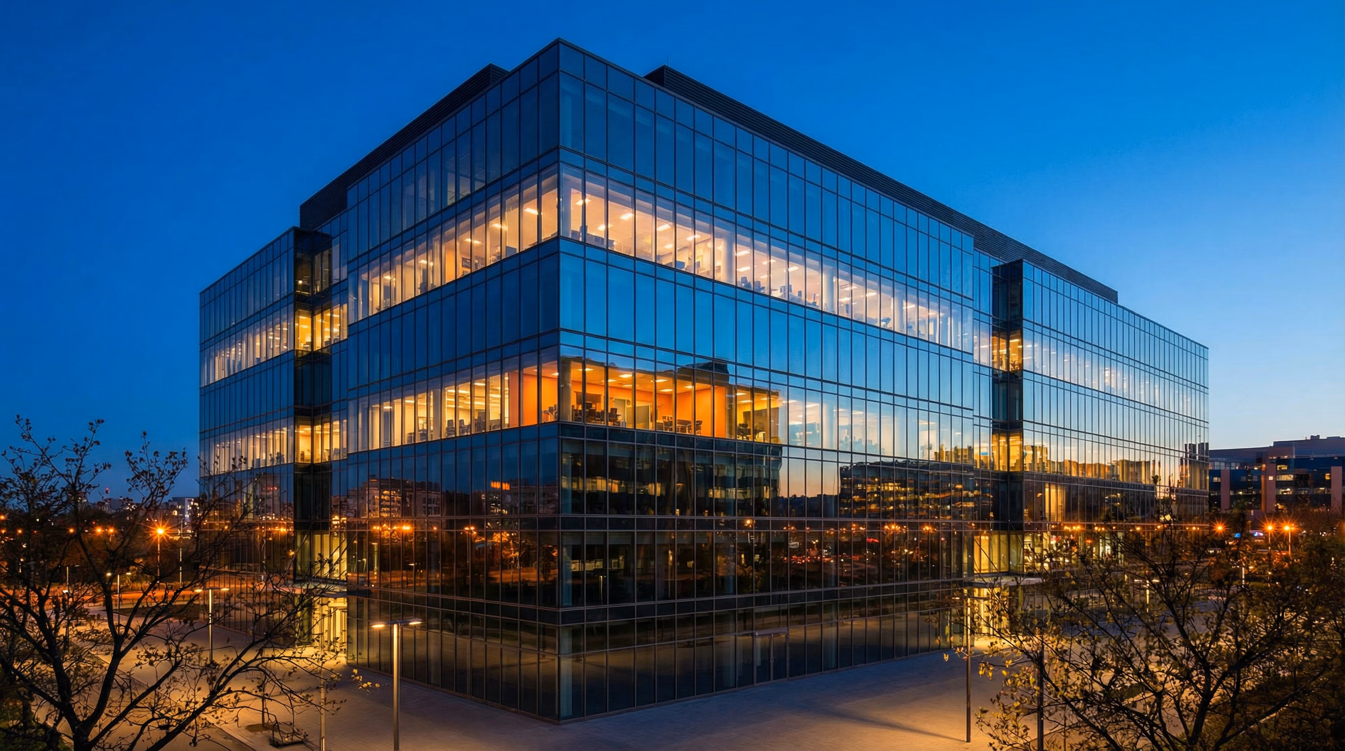 Modern glass-curtain-wall office building lit from within against a deep blue evening sky.
