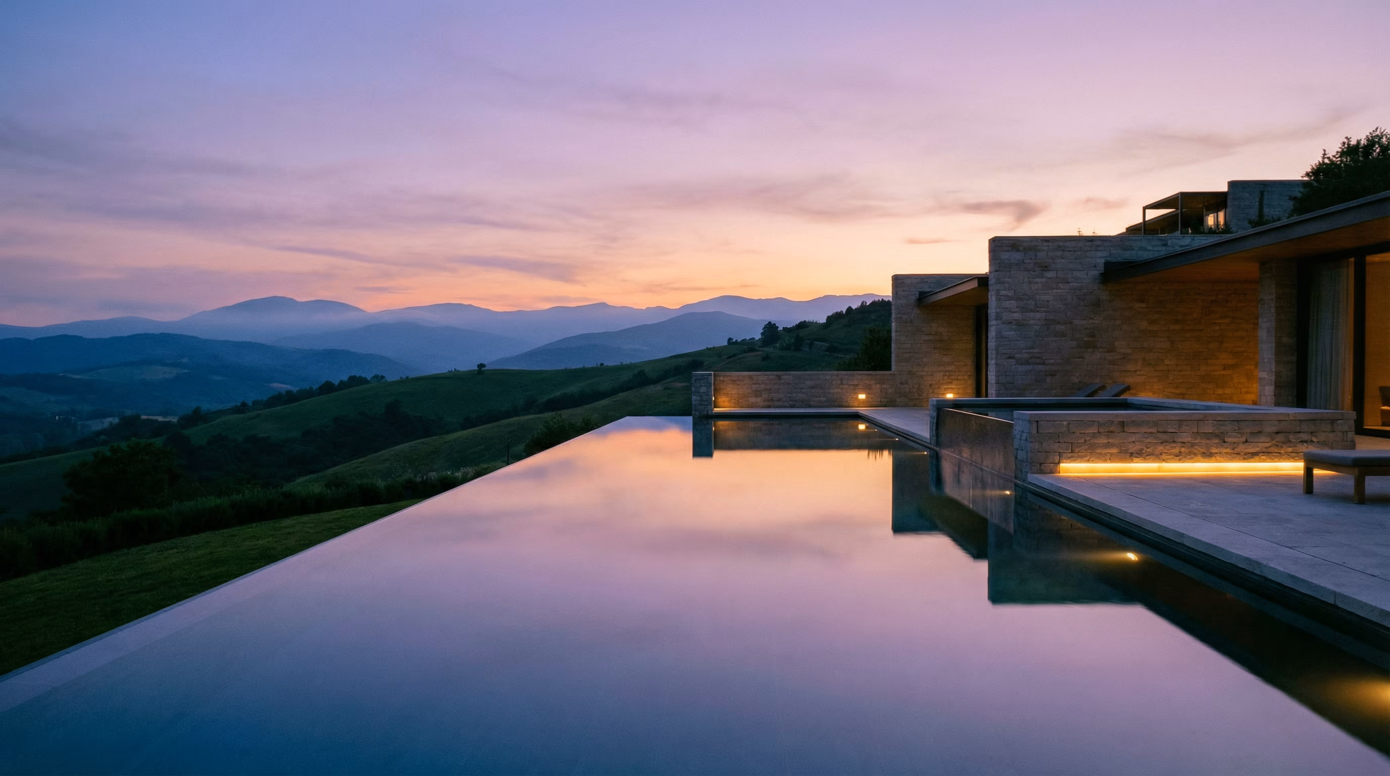 Infinity pool overlooking rolling hills at dusk beside a modern stone building.