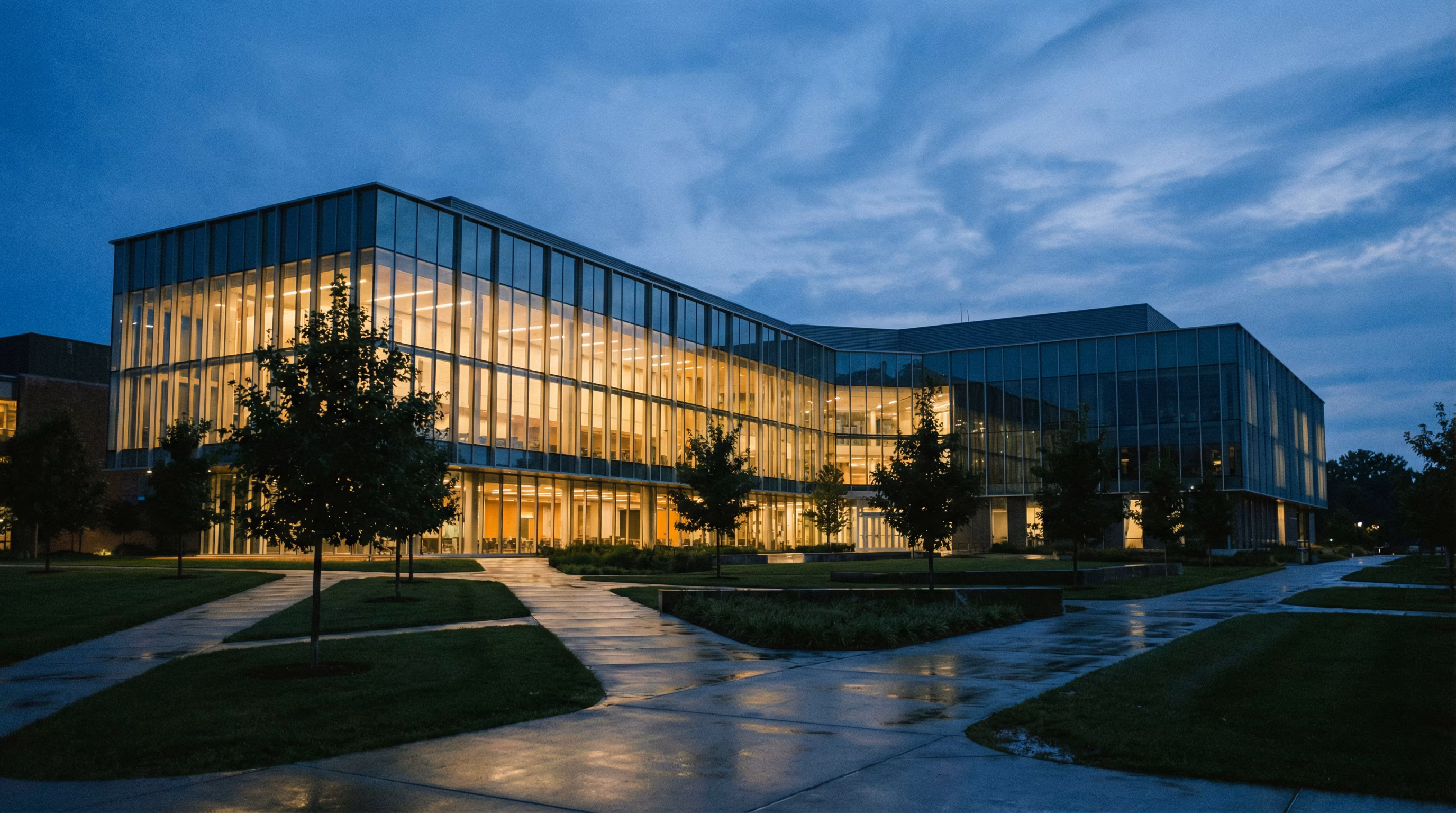 A large modern glass university building illuminated from inside at dusk with pathways and trees in the foreground.