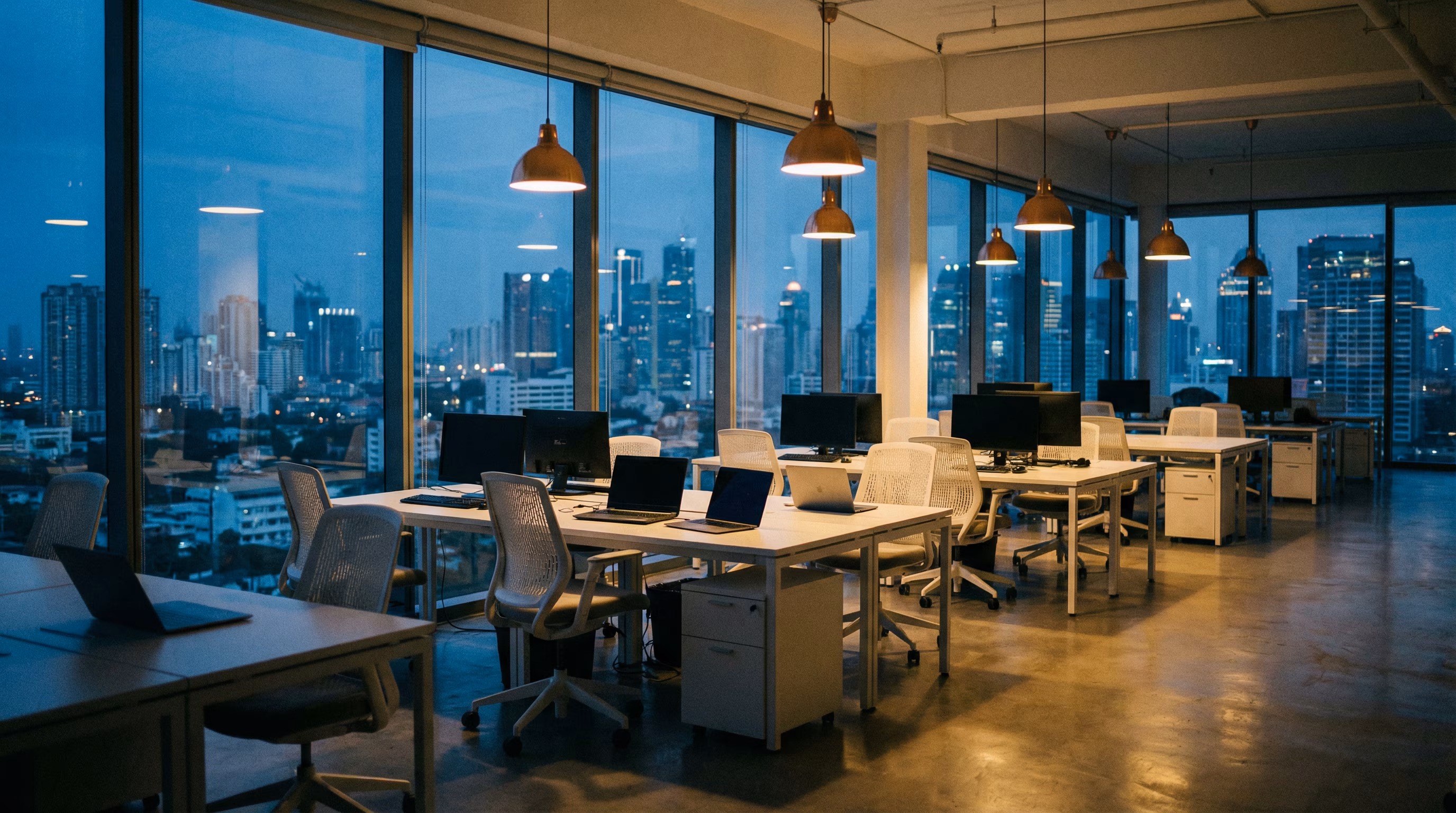 Open-plan office with rows of desks and computers lit by pendant lights against a city skyline at dusk.