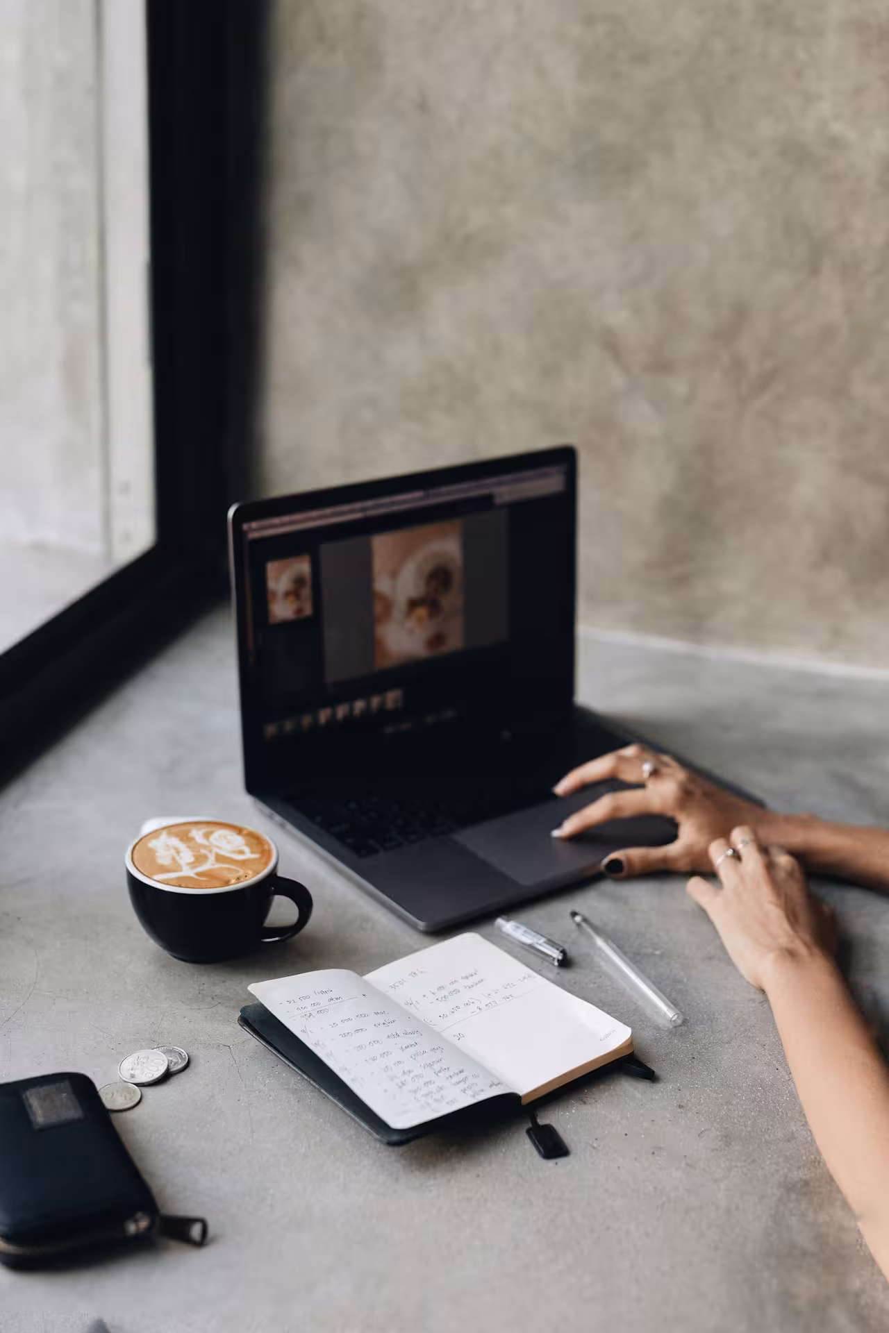 Person typing on a laptop beside an open notebook, a latte, and a small wallet on a concrete surface.