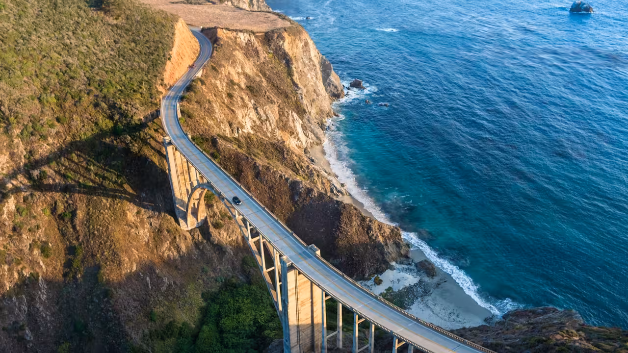 Aerial view of a curved concrete bridge hugging a rocky coastal cliff above bright turquoise water.