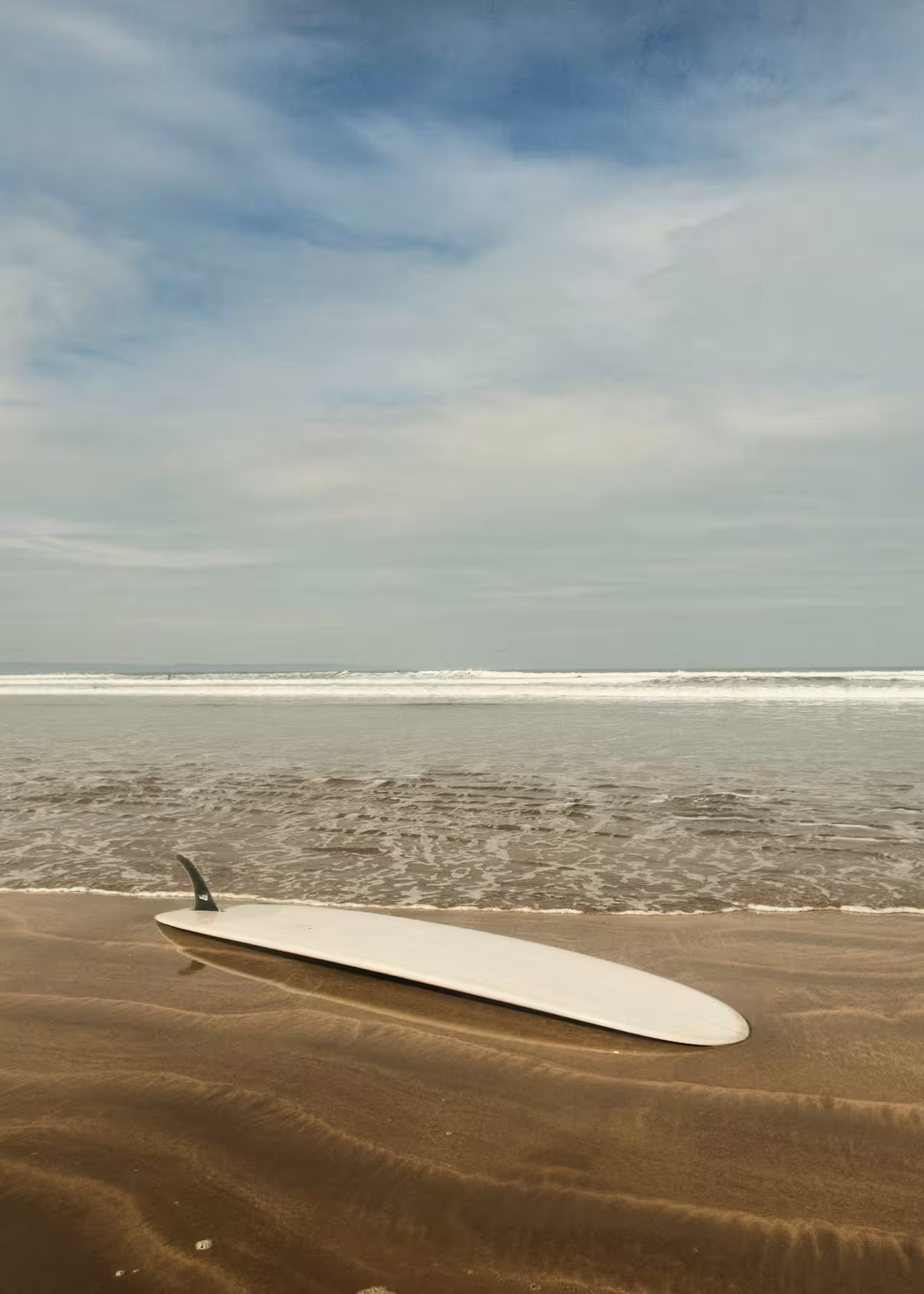 A white surfboard rests fin-up on wet sand at the shoreline with ocean waves in the background.