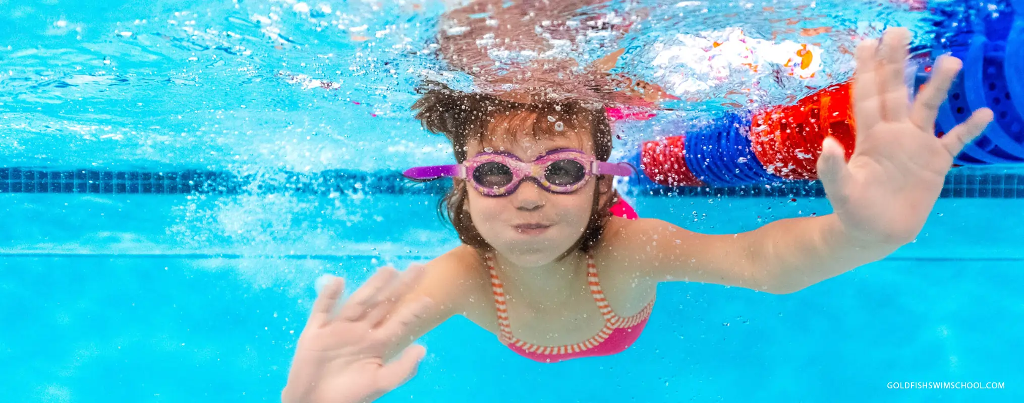 Young girl in pink goggles swimming underwater in a bright pool near lane dividers.