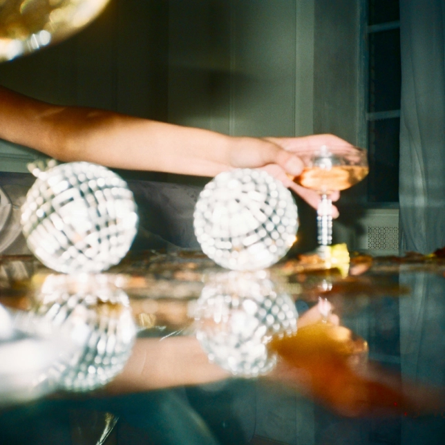 Hand holding a glass of champagne above a reflective surface with decorative disco balls nearby.
