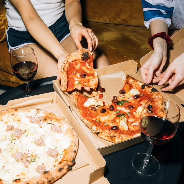 Two people sharing pizzas and glasses of red wine on a table beside a couch.