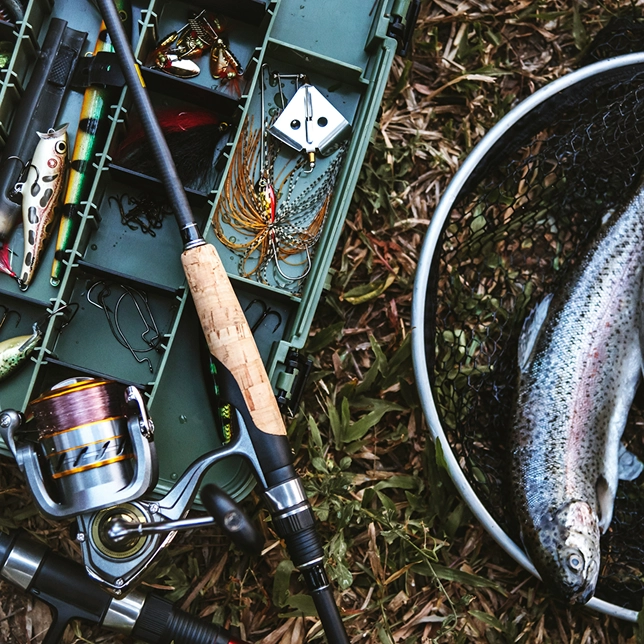Fishing gear including rod, reel, tackle box with lures, and a trout in a net on grass.