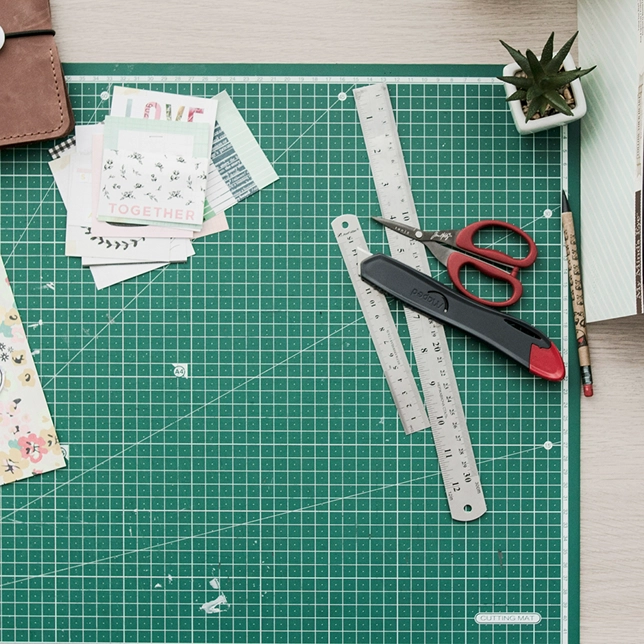 Green cutting mat with rulers, scissors, craft knife, pencil, small plant, and paper scraps on a desk.