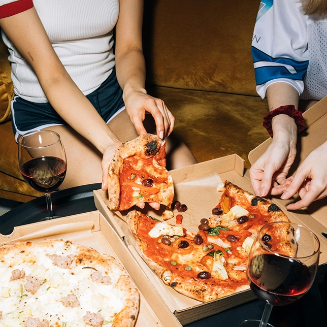 Two people sharing pizza slices from open boxes with glasses of red wine on a black table.