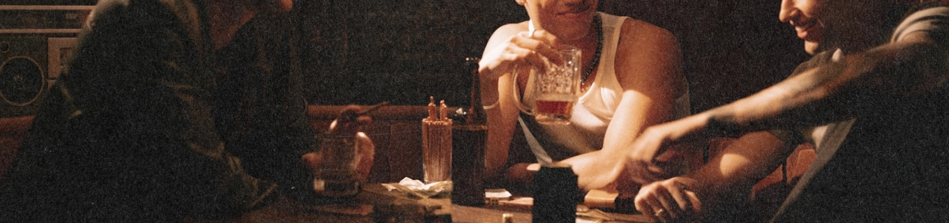 Three men sitting at a table holding drinks and chatting in a dimly lit room.