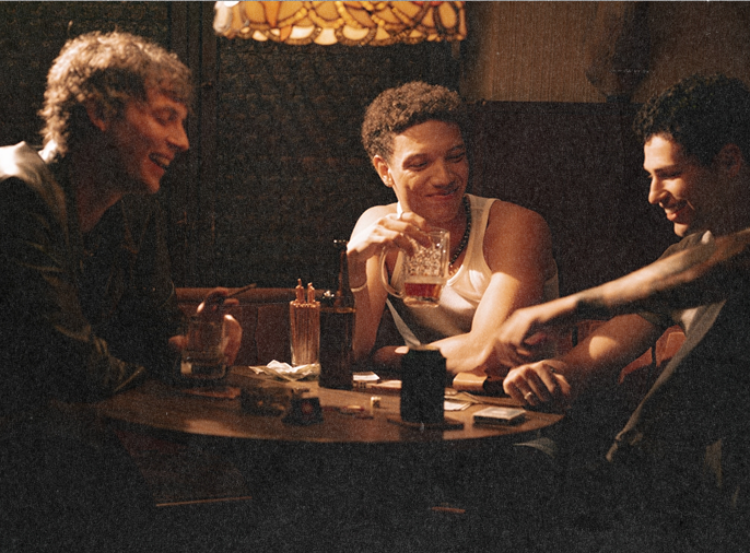 Three young men sitting around a small wooden table in a dimly lit room, smiling and enjoying drinks, cards, and snacks.