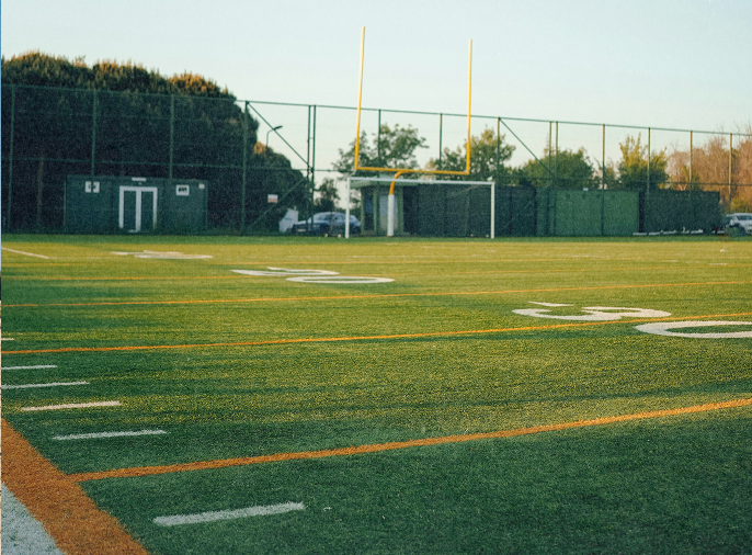 Empty American football field with green artificial turf, white yard lines, and yellow goalposts under clear sky.