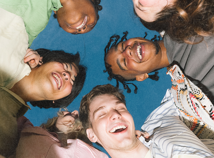 Six diverse young adults standing in a circle looking down at the camera, smiling and laughing against a blue sky background.
