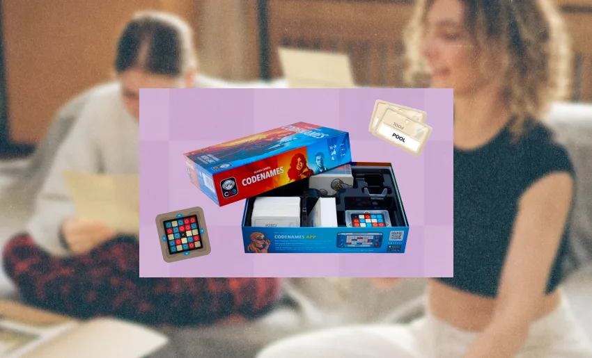 Open box of the Codenames board game showing the game cards, timer, and a rules card on a purple background, with two blurred people in the background holding cards.