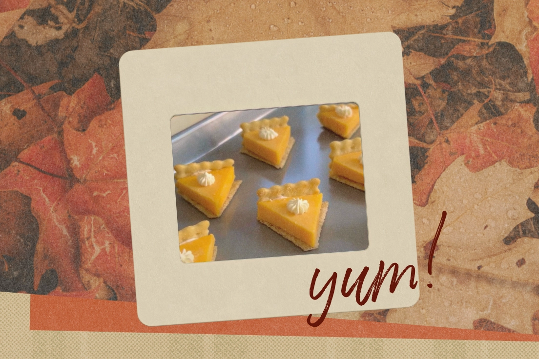 Close-up of triangular mini pumpkin tarts with a dollop of whipped cream on top, arranged on a metal tray with an autumn leaf background and the word 'yum!' in brown script.