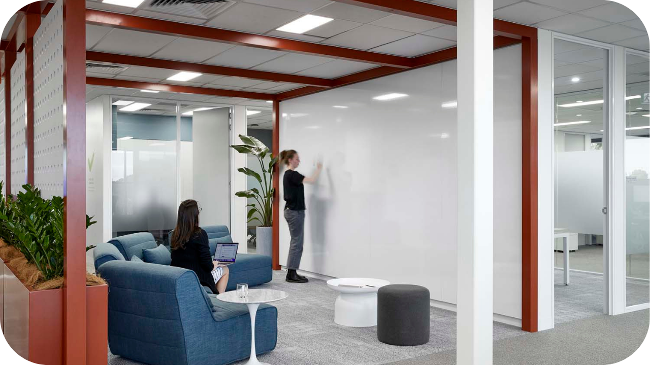 Collaborative office fit out with lounge seating, greenery, and a woman writing on a large whiteboard wall during a meeting.