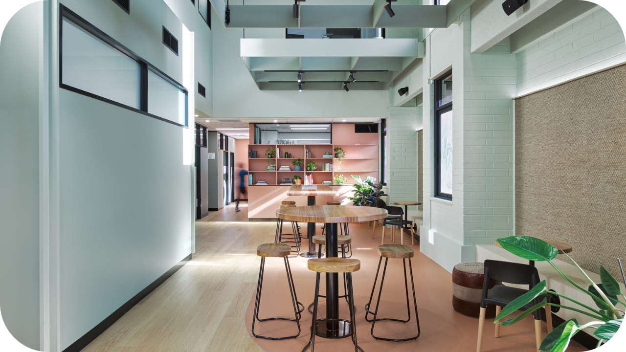 Open office breakout area with round timber tables, high stools, and pastel joinery shelving.